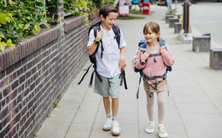 Två barn med ryggsäckar och smartklockor går och skrattar på en trottoar på väg till skolan. Båda bär Xplora barnsmartklockor för säkerhet och kommunikation.
