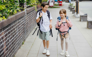 Två barn med ryggsäckar och smartklockor går och skrattar på en trottoar på väg till skolan. Båda bär Xplora barnsmartklockor för säkerhet och kommunikation.