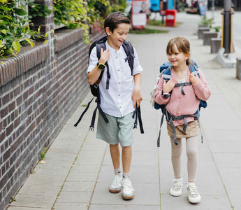 Två barn med ryggsäckar och smartklockor går och skrattar på en trottoar på väg till skolan. Båda bär Xplora barnsmartklockor för säkerhet och kommunikation.