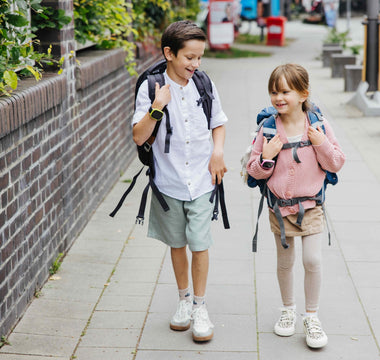 Två barn med ryggsäckar och smartklockor går och skrattar på en trottoar på väg till skolan. Båda bär Xplora barnsmartklockor för säkerhet och kommunikation.