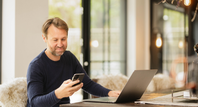 En man med skägg och mörkblå tröja sitter vid ett bord i en modern, ljus inomhusmiljö. Han håller en mobiltelefon i handen och tittar på den medan han arbetar på en bärbar dator. Bakgrunden har stora fönster och varm belysning.