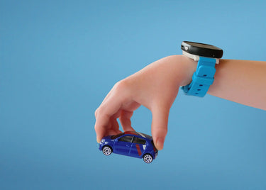 Ett barns hand med en blå smartklocka håller en liten blå leksaksbil mot en enfärgad ljusblå bakgrund. Smartklockan har en svart rektangulär skärm och ett starkt blått armband, vilket kontrasterar mot barnets ljusa hud.