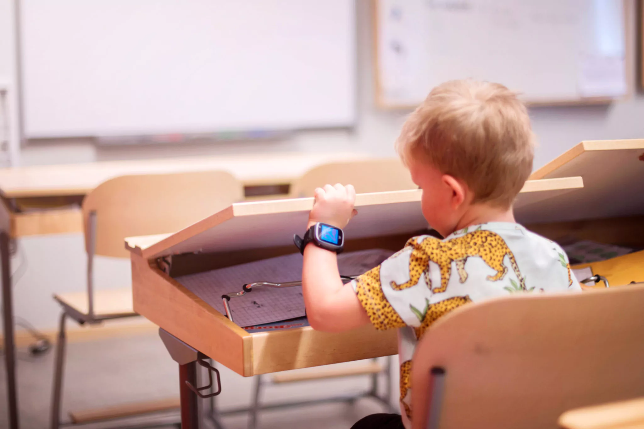 en pojke sitter i skolan vid sin skolbänk md en blå xplora klocka på vänster arm. Klockan är i skolläge